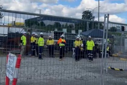 Concrete trucks and traides in high vis ppe blocked behind fence by union members