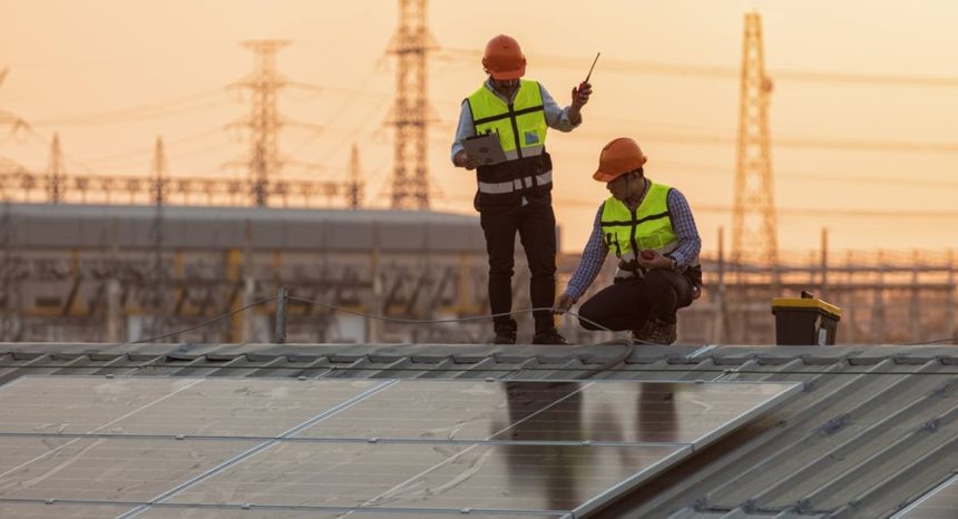 Men in high vis and hard hats standing on rooftop with solar panels with electrical towers in the background