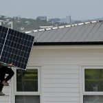 Man on ladder taking solar panel up to roof of home