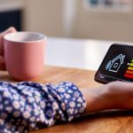 Woman drinking cup of tea at table looking down at phone displaying home energy readout