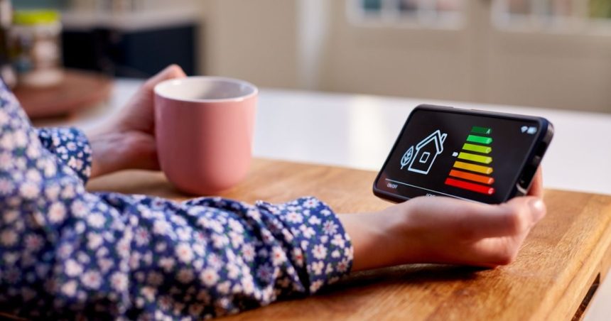 Woman drinking cup of tea at table looking down at phone displaying home energy readout