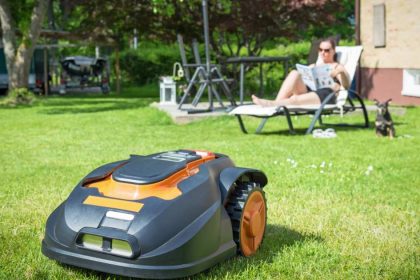 Robotic mower on lawn in front on woman in lawn chair reading magazine next to dog