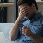 man holding credit card with palm over his face in front of laptop on couch