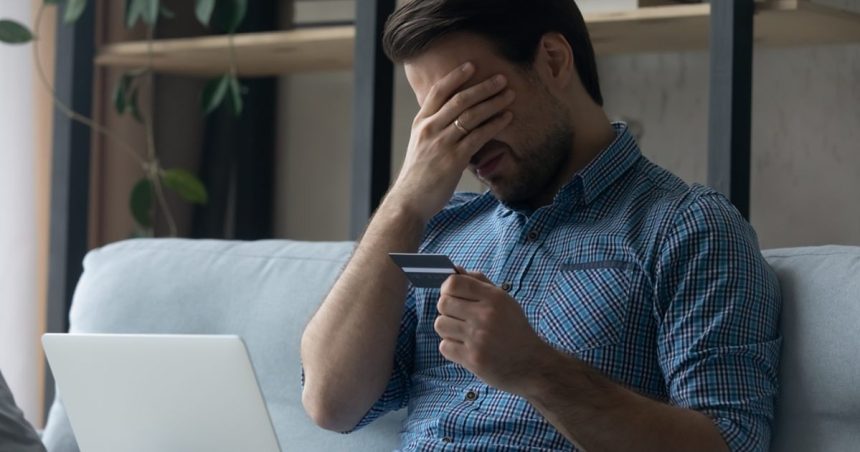 man holding credit card with palm over his face in front of laptop on couch