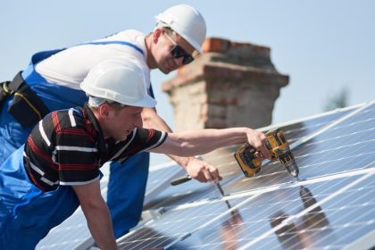 Two men n overalls and hard hats on rood installing solar panels